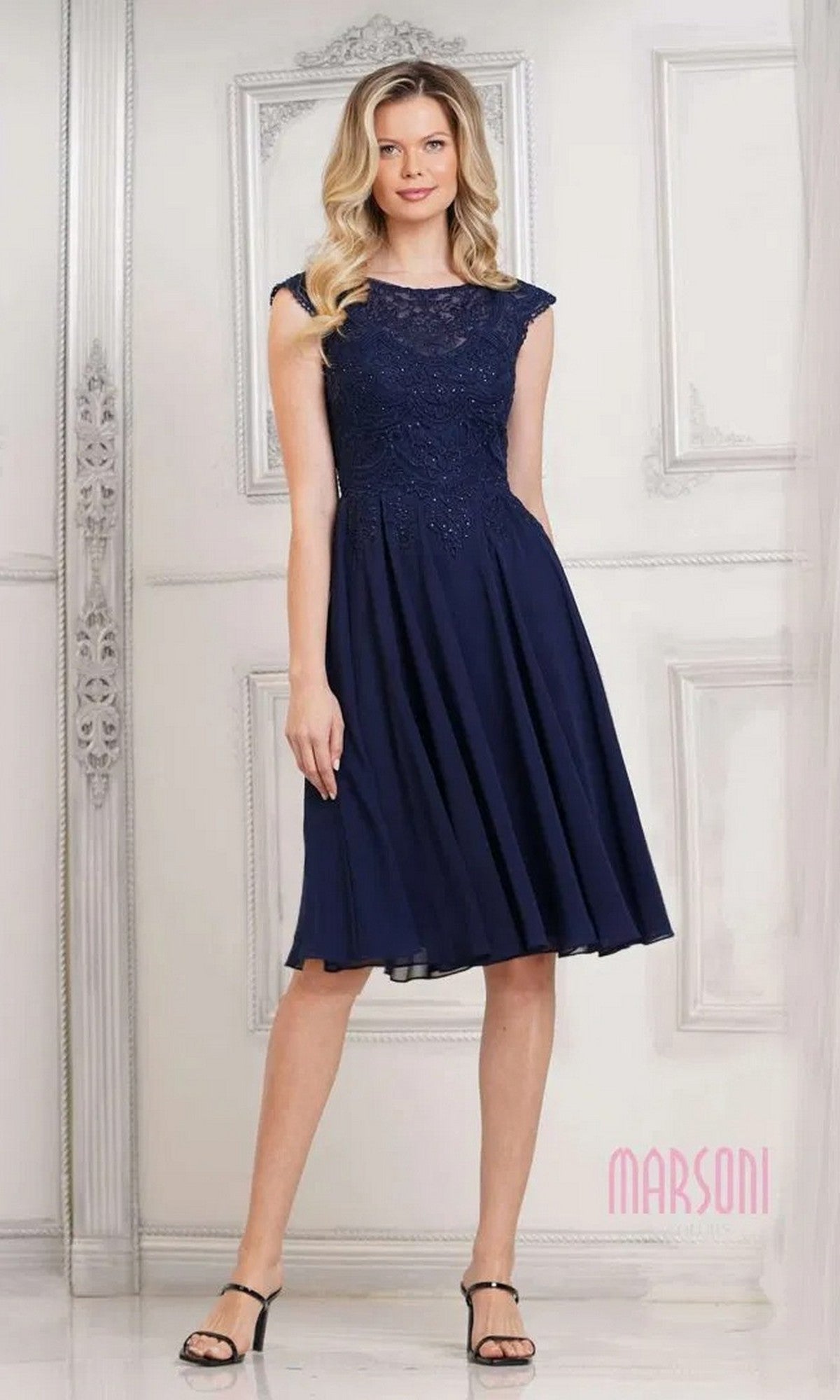 A woman stands indoors wearing the Marsoni M238S knee-length semi-formal dress in navy, featuring a lace-embroidered bodice, chiffon skirt, cap sleeves, and black strappy heels.